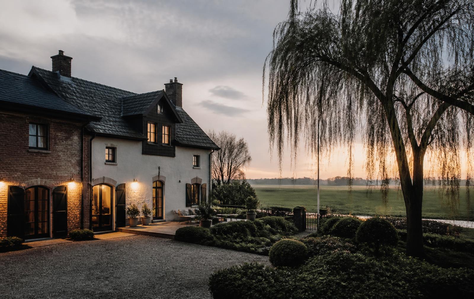 Landhaus aus Backstein und weißem Putz mit warm erleuchteten Fenstern am Rand der Rheinwiesen in der Dämmerung